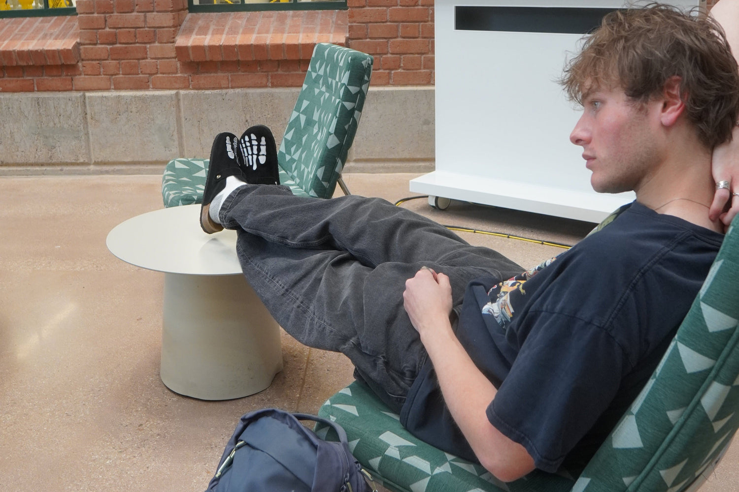 Person lying on a green cushioned chair with a backpack next to them, outside a building.

